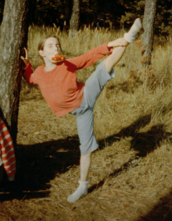 Maria Lichtenberg praktiziert bereits als kleines Mädchen spielerisch Yoga und hebt in der freien Natur ein Bein hoch in die Luft. Sie lehnt an einem Baum. Ihr Gesicht ist verschmitzt.