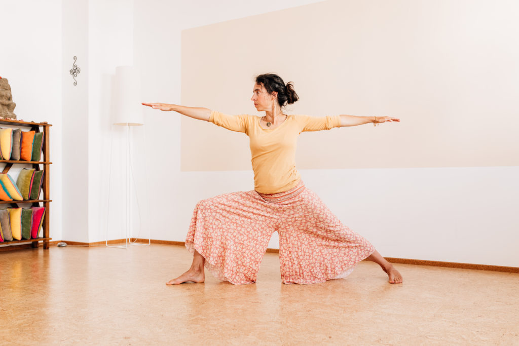 Die Yogalehrerin Maria Lichtenberg verweilt fest/ stabil und gleichzeitig entspannt und fokussiert in der Kriegervariante 2/ Vīrabhadrāsana 2 in ihrem Yogaraum in Leipzig.