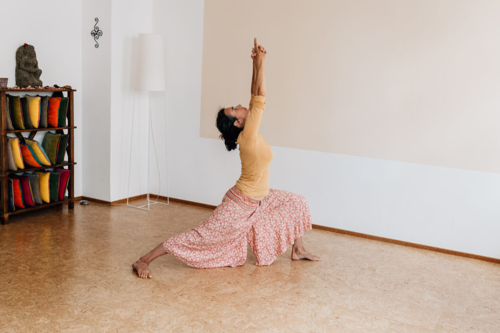 Die Yogalehrerin Maria Lichtenberg verweilt fest/ stabil und gleichzeitig entspannt in der Kriegervariante / Vīrabhadrāsana 1 in ihrem Yogaraum in Leipzig.