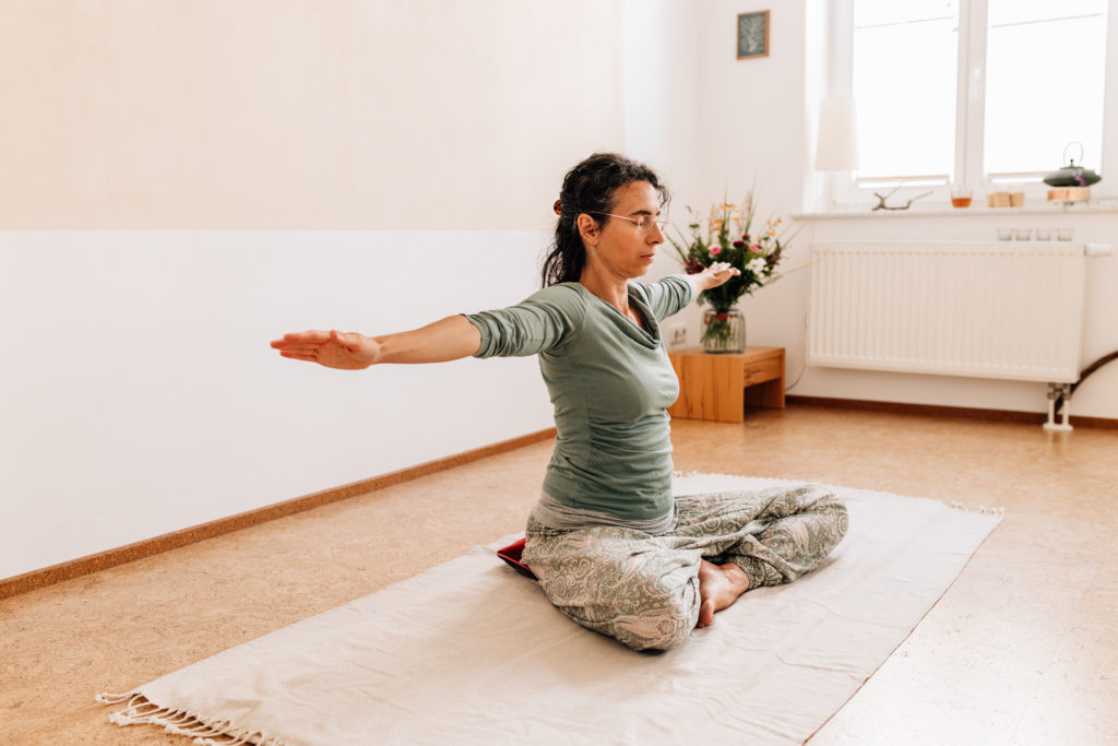 Die Yogalehrerin Maria Lichtenberg breitet ihre Arme seitlich auf Schulterhöhe aus und verlängert sie zu den Seiten hin in ihrem Yogaraum in Leipzig.
