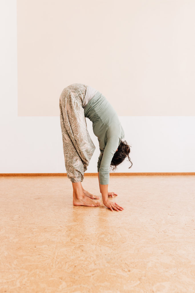 Die Yogalehrerin Maria Lichtenberg verweilt in einer Vorbeuge im Stehen - der Körper hängt entspannt nach vorn unten, die Arme berühren den Boden in ihrem Yogaraum in Leipzig.