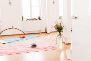Ein Blick in den lichtvollen Yogaraum der Yogalehrerin Maria Lichtenberg mit den ausgelegten Yogamatten aus Schafschurwolle, darauf farbige Baumwolldecken und kleine Meditationskissen. Ein Blumenstrauß und Kerzen schaffen eine angenehme Atmosphäre.
