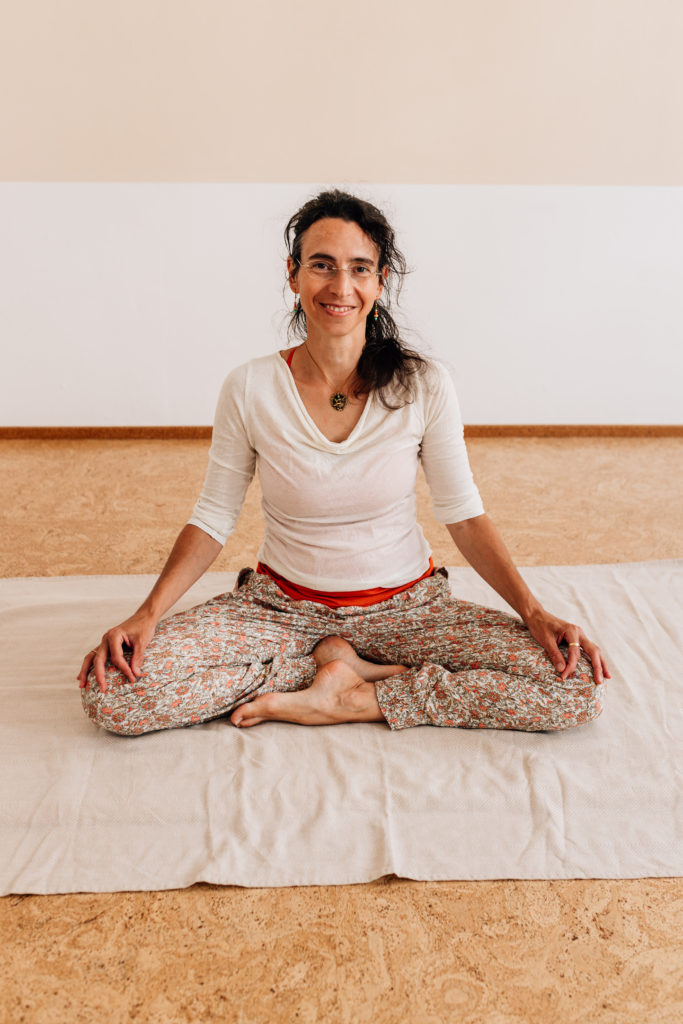 Maria Lichtenberg lächelt in ihrem Yogaraum in Leipzig in einer entspannten meditativen Sitzhaltung freudig in die Kamera.