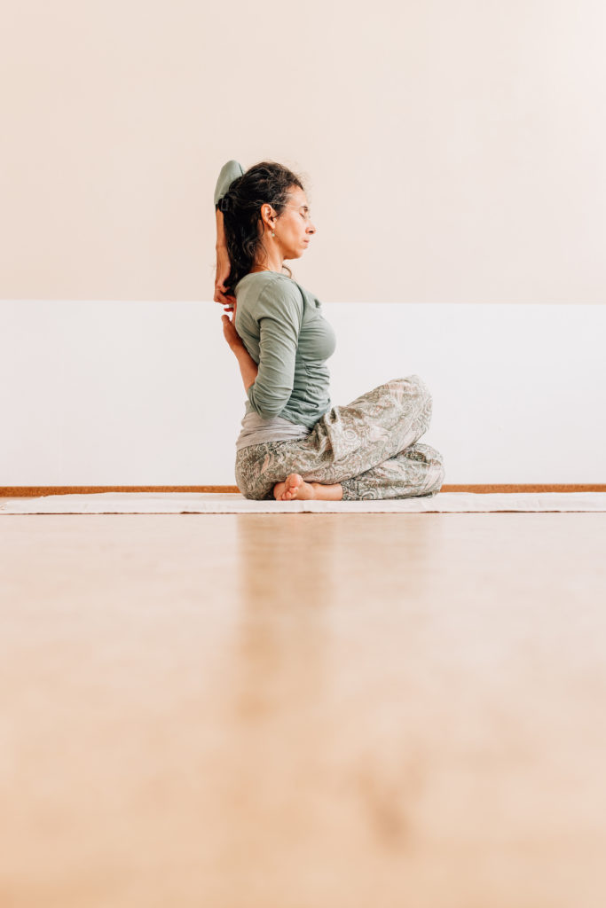 Die Yogalehrerin Maria Lichtenberg hält hinter ihrem Rücken mit der einen Hand die andere während die Oberschenkel - insbesondere die Knie - übereinander liegen in ihrem Yogaraum in Leipzig.
