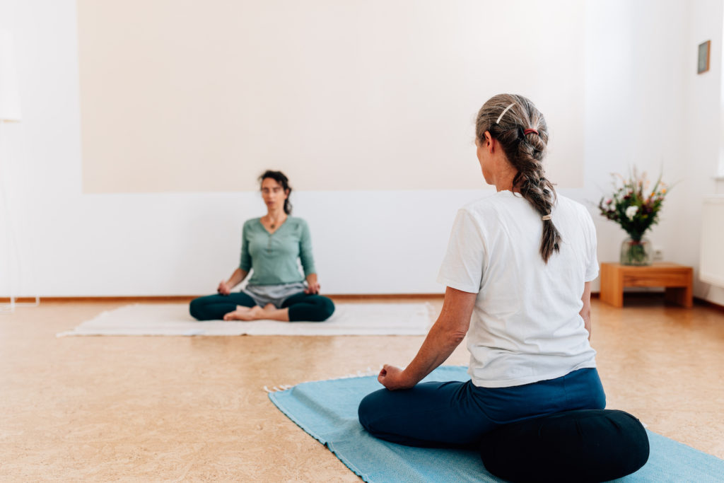 Die Yogalehrerin Maria Lichtenberg beginnt mit ihrer Yogaklasse (eine Schülerin ist sichtbar) die Unterrichtsstunde in der meditativen Sitzhaltung mit einer Atembetrachtung. Die Wirbelsäule richtet sich von innen heraus auf, die Schultern und das Steißbein sinken nach unten, das Brustbein hebt sich und unnötige Anspannung kann nach und nach weichen.